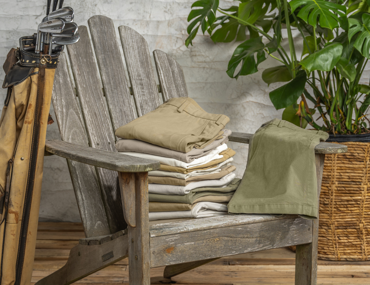 A stack of Bills Khakis shorts in various colors sit on a weather outdoor chair with a vintage golf bag full of golf clubs leaning on it. Greenery sits in the background