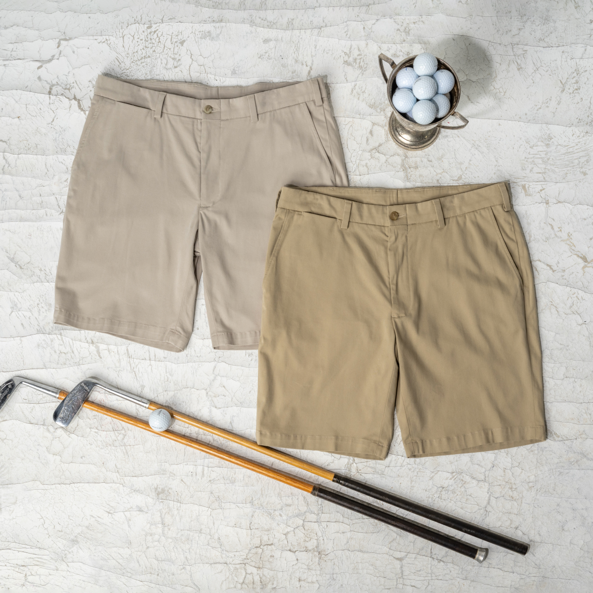 Two pairs of Bills Khakis Travel Twill shorts lay on a weathered surface alongside vintage golf clubs and some golf balls
