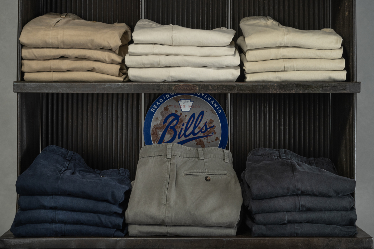 An industrial piece of shelving with a rustic metal Bills Khakis logo sign is stocked with five colors of Bills Khakis Heritage Wash Pants