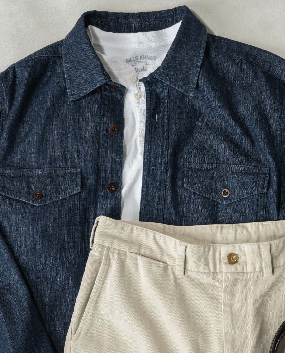 A cropped in, laydown shot of a Bills Khakis Lightweight Denim Overshirt over a Bills Henly along with Bills bone colored khaki pants
