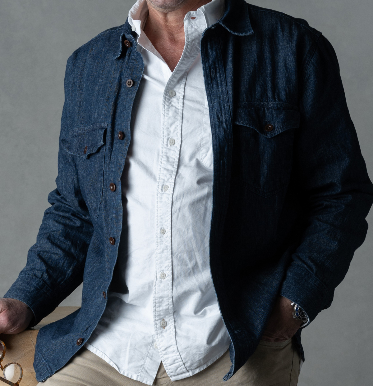 A man posing in front of a grey backdrop. You cannot see his face. He is wearing the Bills Khakis Lightweight Denim Overshirt over a Bills white Standard Fit shirt. He is wearing Bills Khaki pants and has a watch on and is carrying glasses