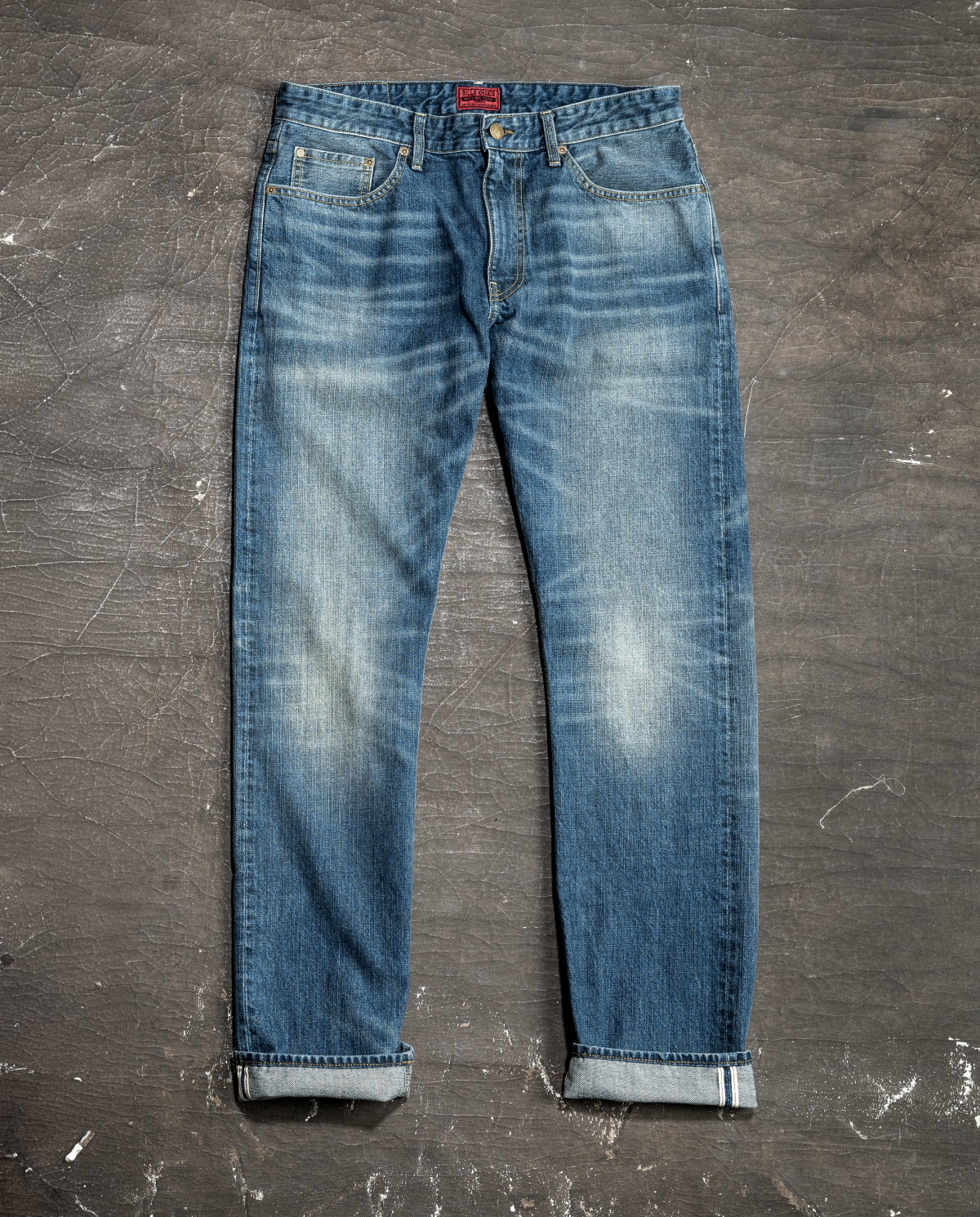 A pair of Bills Khakis Red Selvage Denim pants lay flat on a grey industrial background