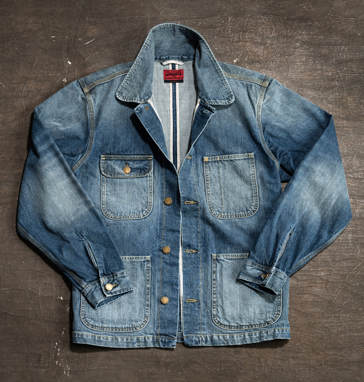 A Bills Khakis Red Selvage Denim jacket lays flat on a grey industrial background