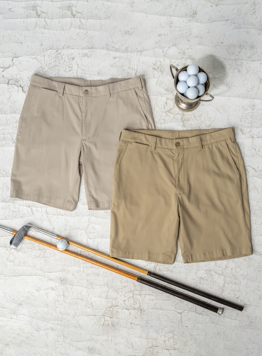 Two pairs of Bills Khakis Travel Twill shorts lay on a weathered white surface alongside vintage golf equipment and memorabilia