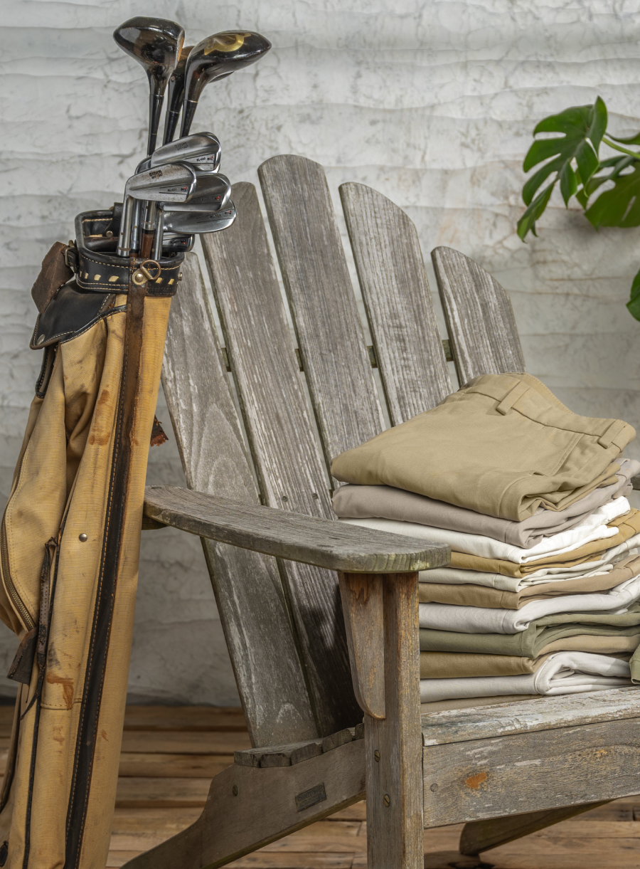 Pairs of Bills Khakis bottoms sit folded on a weathered outdoor chair alongside vintage golf clubs + bag