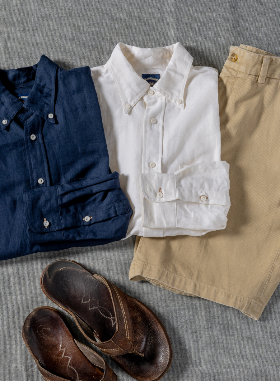 Two Bills Khakis shirts and a pair of shirts lay on a grey surface alongside a pair of sandals