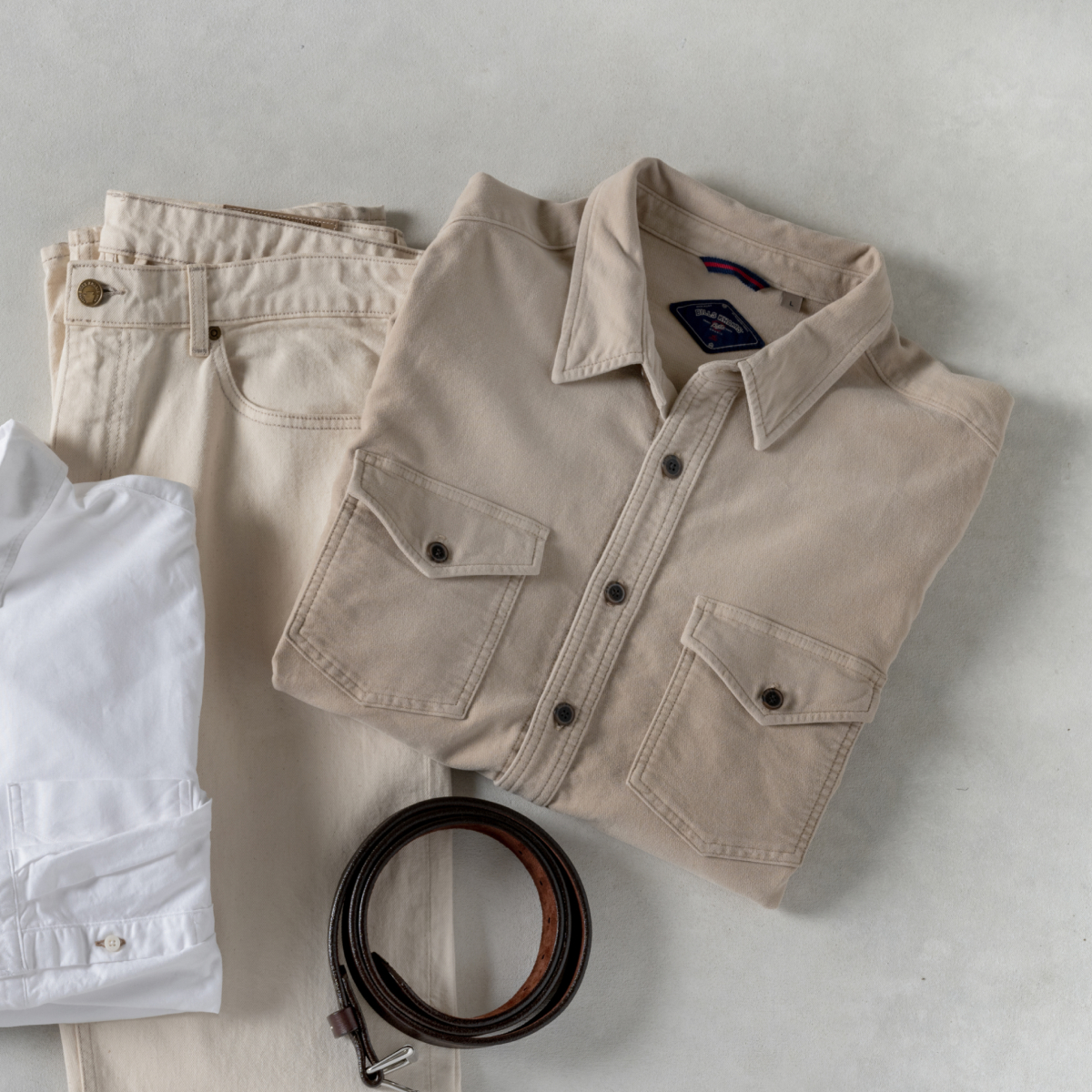 Light shades of Bills Khakis - an overshirt, linen shirt, pants, belt - lay on a light colored surface