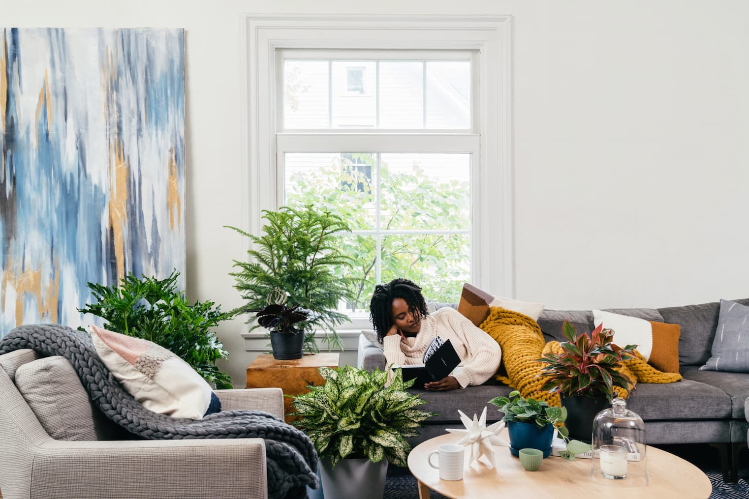 woman reading book on sofa, surrounded by plants