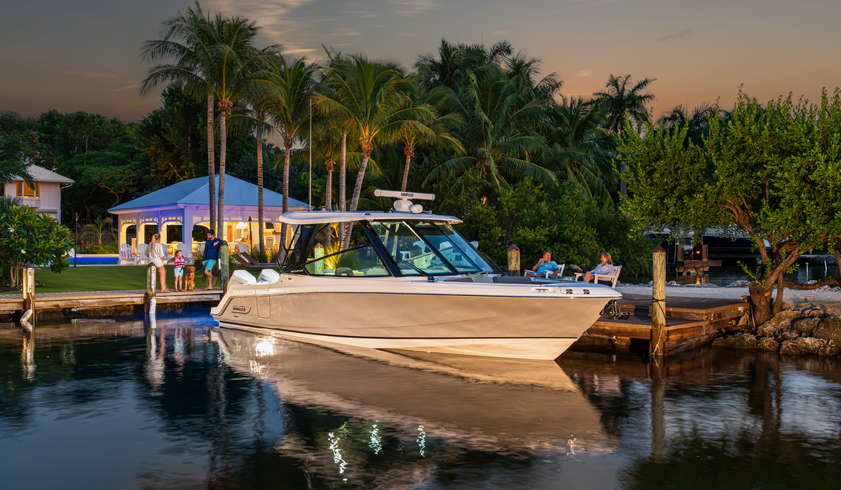 A sleek, modern powerboat sits docked as dusk settles, reflecting in the calm water. A family waits on the pier, while another couple relaxes nearby.