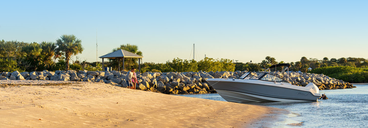 A gray and white Boston Whaler boat resting on a sunlit sandy shore, with a line of large rocks separating the beach from a green landscape featuring palm trees and a small white gazebo.