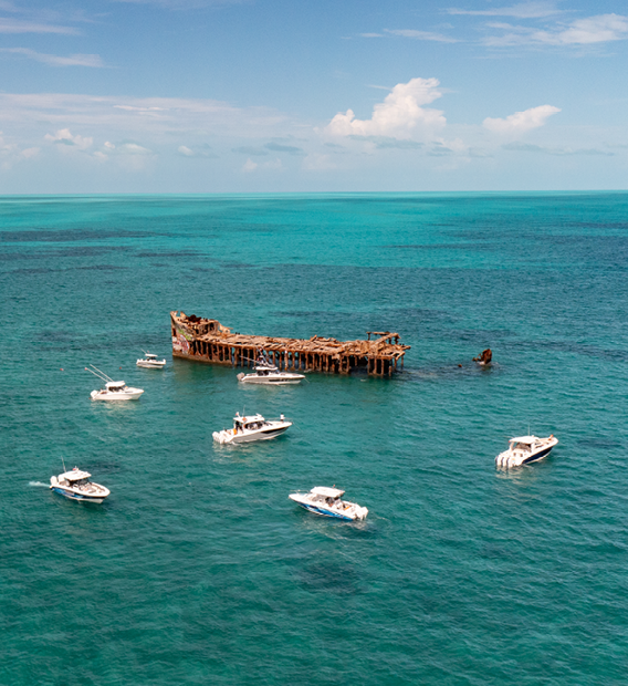 Aerial view of a large, rusty shipwreck in clear turquoise water, surrounded by several white Boston Whaler under a partly cloudy sky.