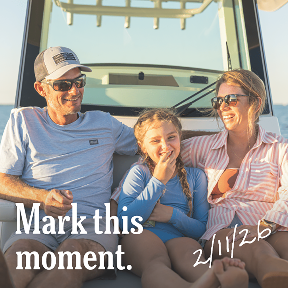 A family of three (father, mother, and young daughter) smiling and laughing together on a boat, with the text "Mark this moment." and "2/11/26" overlaid.