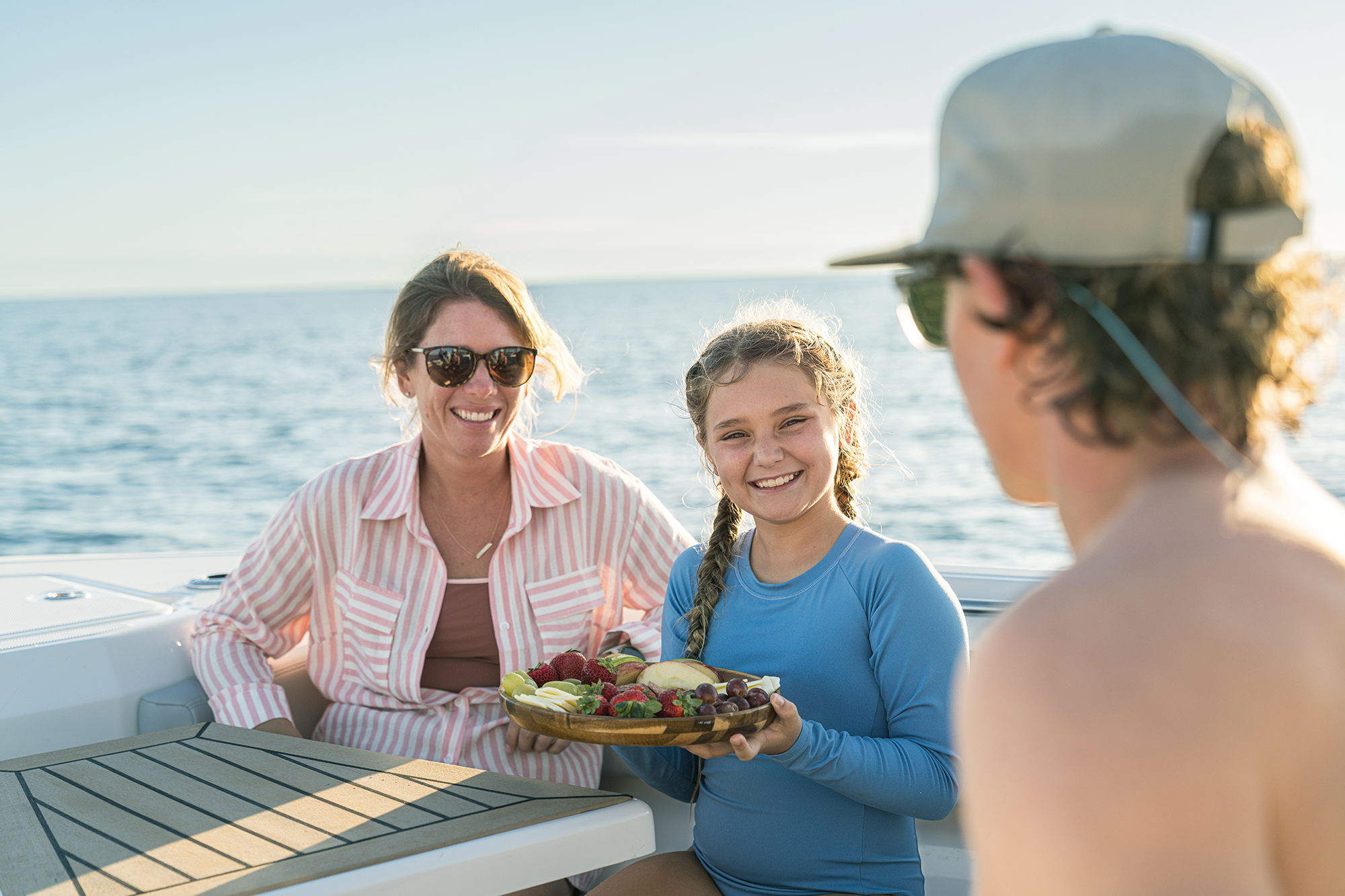 A smiling woman and girl on a boat, with the girl holding a platter of fresh fruit, with the ocean in the background.