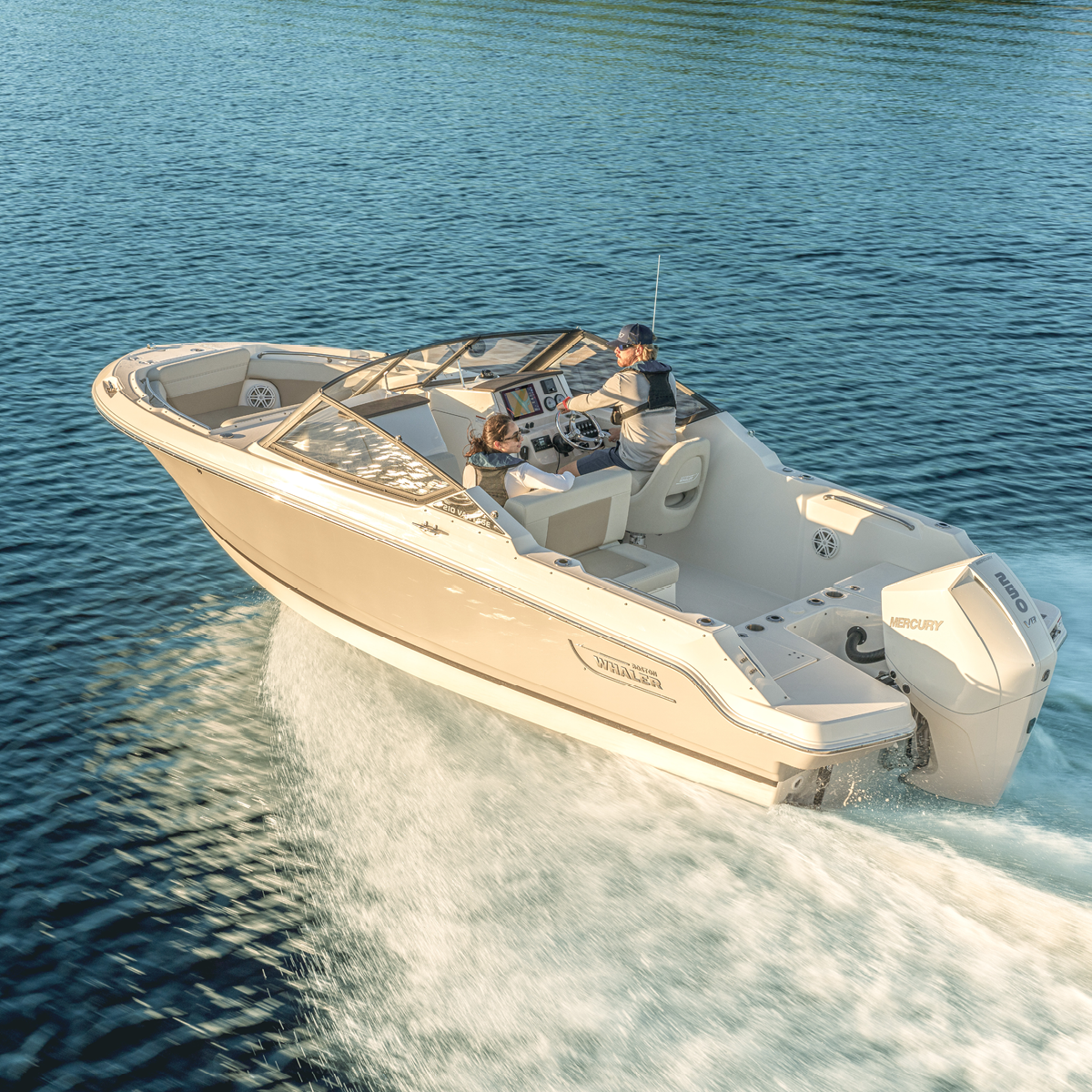 A light-colored Boston Whaler boat with an adult and child aboard, speeding across blue water and creating a white wake.