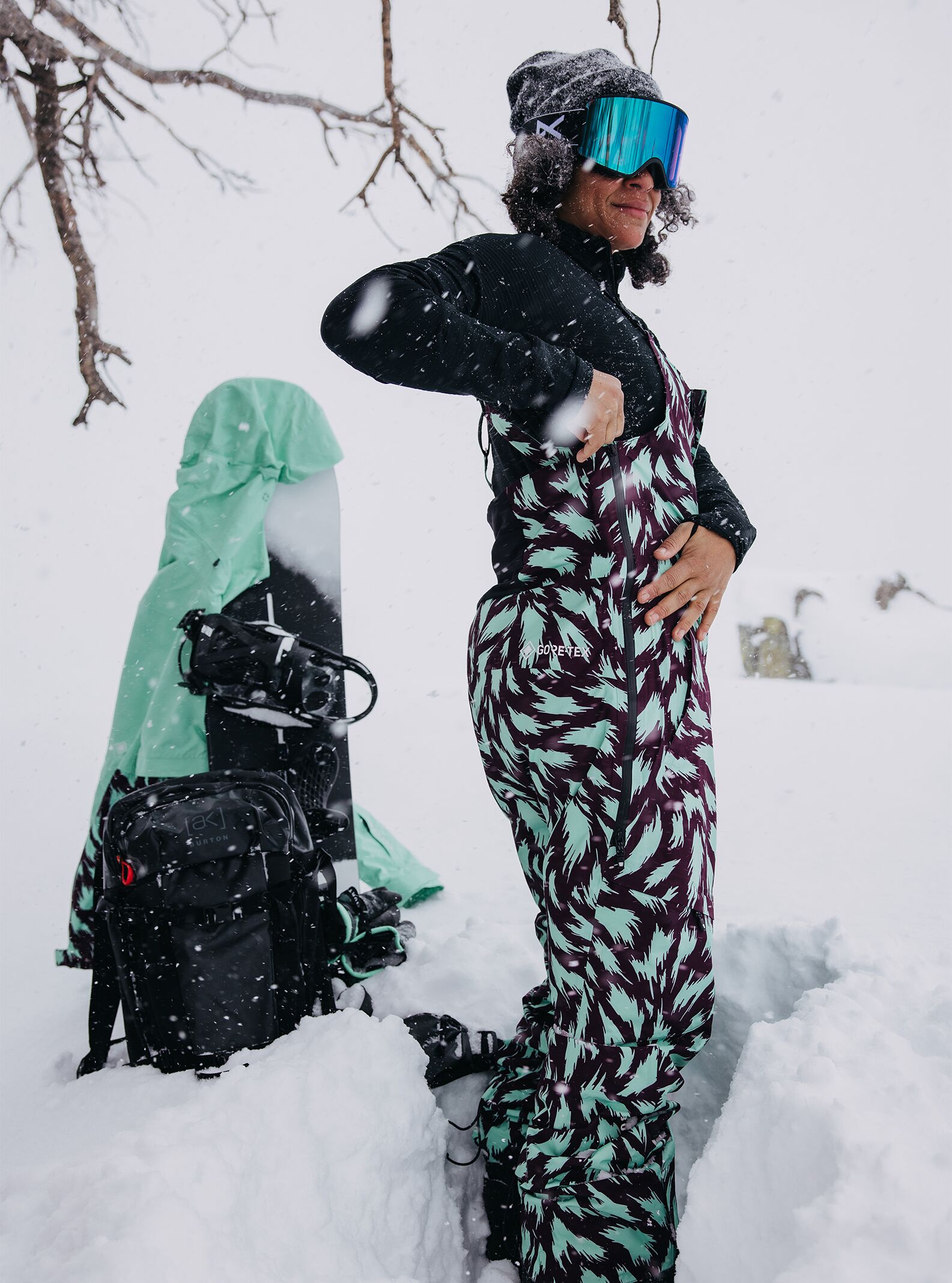 Women's Burton [ak] Kimmy GORE-TEX 2L Bib Pants in a teal and purple fur pattern on a model in the deeo snow with a Burton Jacket and Snowboard in the background