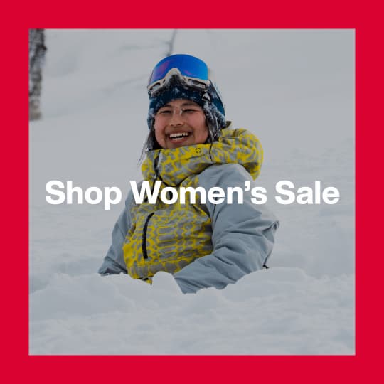 A darkened image of a Burton rider sitting in powder wearing the [ak] Kimmy Anorak in a yellow and gray print in the background. The text 'Shop Women's Sale' is in white on top.