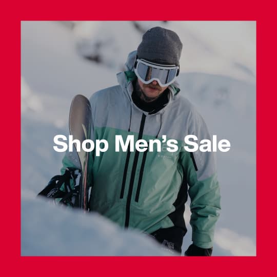A darkened image of a Burton walking in snow carrying a Burton Snowboard wearing the [ak] Swash Jacket in teal and white in the background. The text 'Shop Men's Sale' is in white on top.