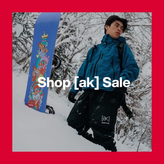 A darkened image of a snowboarder standing in the powder with trees surrounding them wearing Burton [ak] Outerwear in teal and black. The text 'Shop [ak] Sale' is in white on top.