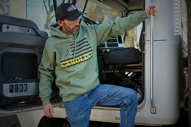 A man wearing the Heritage Stars and Stripes Hoodie in green and an American Flag Trucker hat in black. Shop men's apparel.