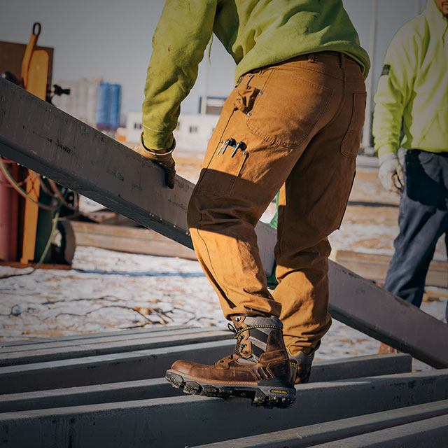 A man wearing the Cancellor Comp Toe Hi boot in brown carrying a metal beam. Shop sale. 