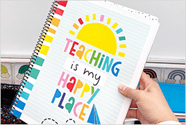 A teacher's hand holding a Happy Place teacher planner with a classroom in the background.