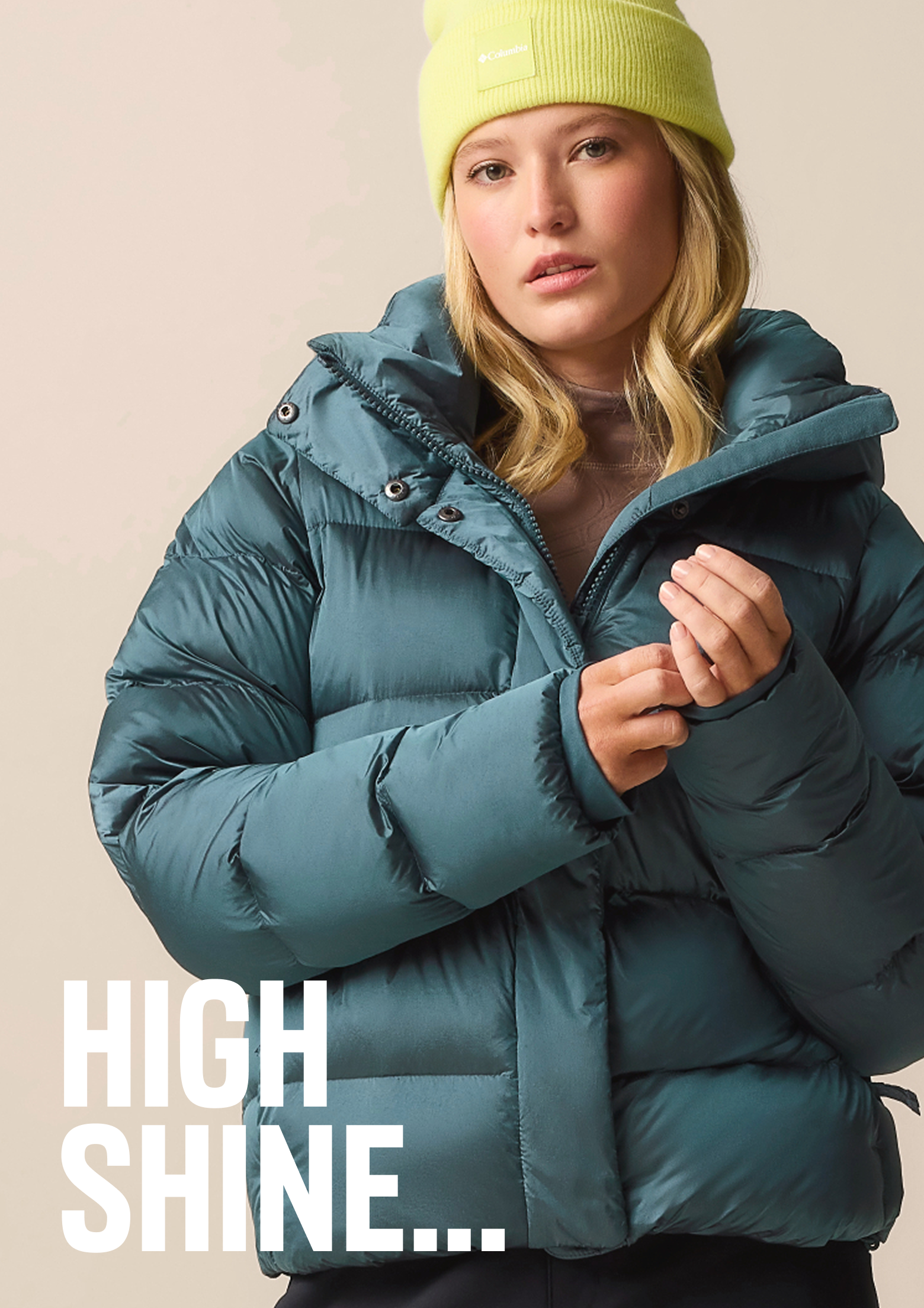 A woman wearing a shiny puffy blue jacket and neon beanie with text: HIGH SHINE