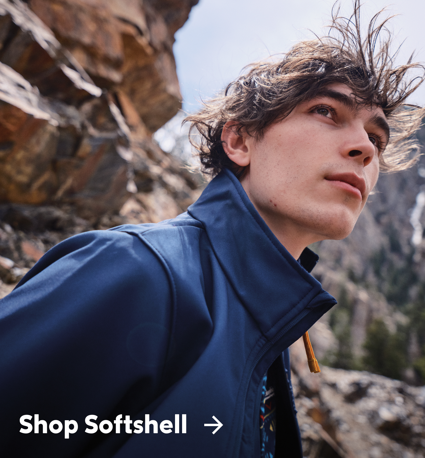 A man wearing a blue jacket in a rocky landscape with text: Shop Softshell