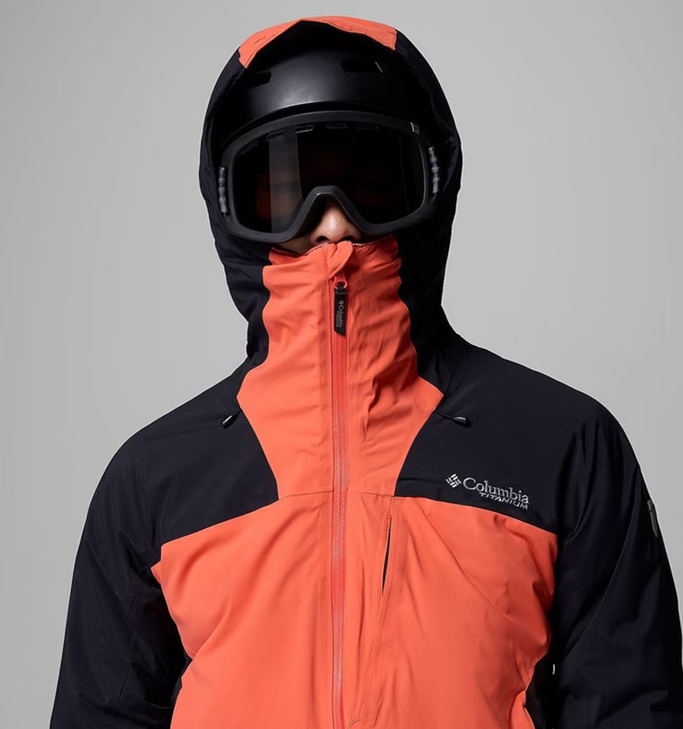 A man wearing an orange and black ski jacket with the hood up and goggles on.