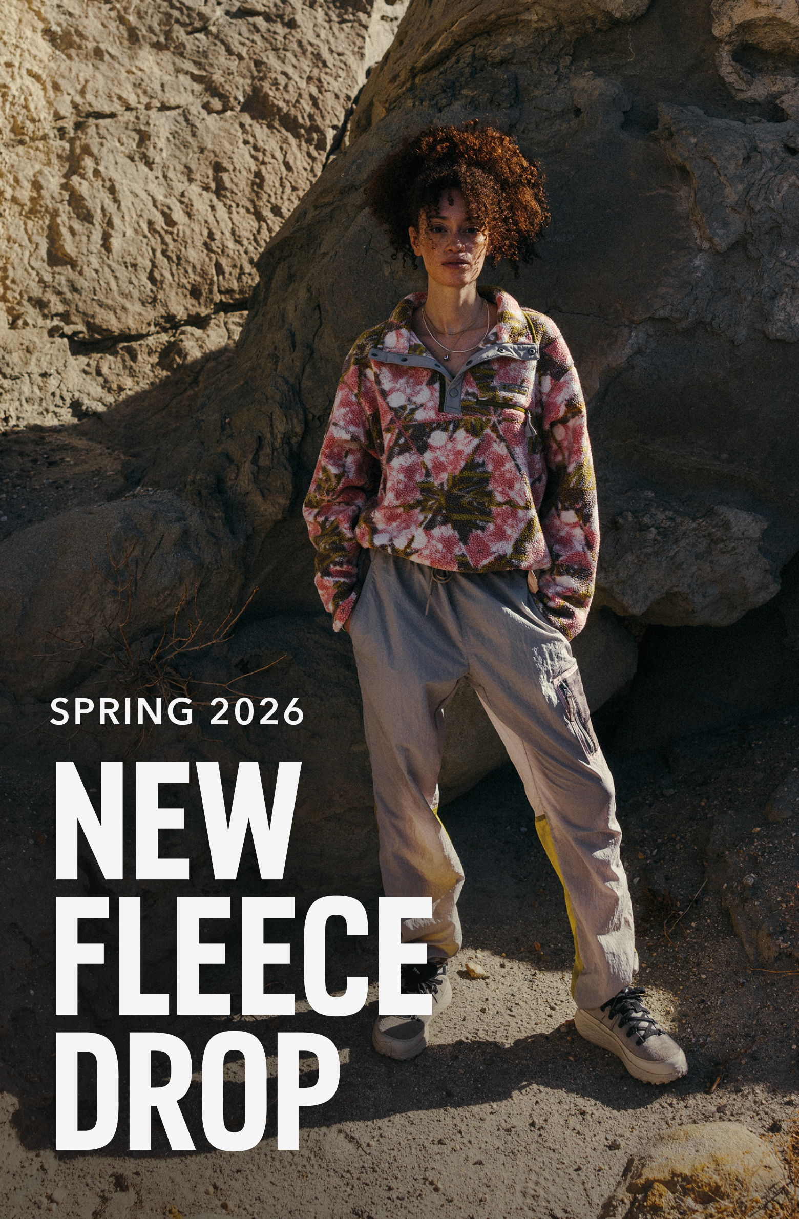 A woman standing next to a rocky wall on a hike wearing a printed fleece, hiking pants and shoes. Text says NEW FLEECE DROP