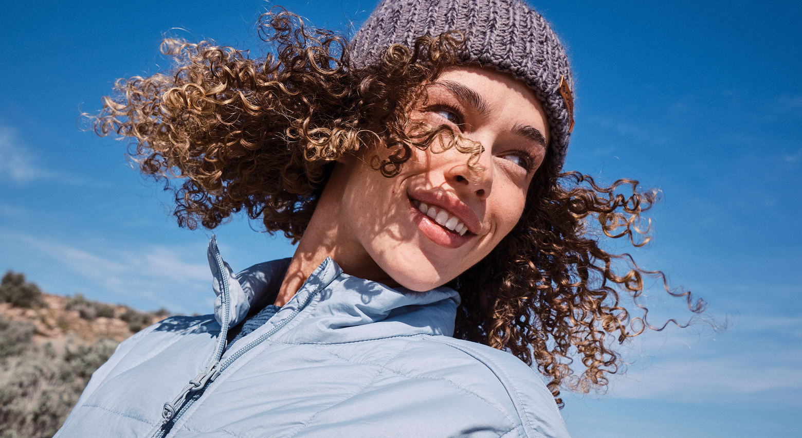 A woman wearing a blue puffer and beanie against a blue sky.