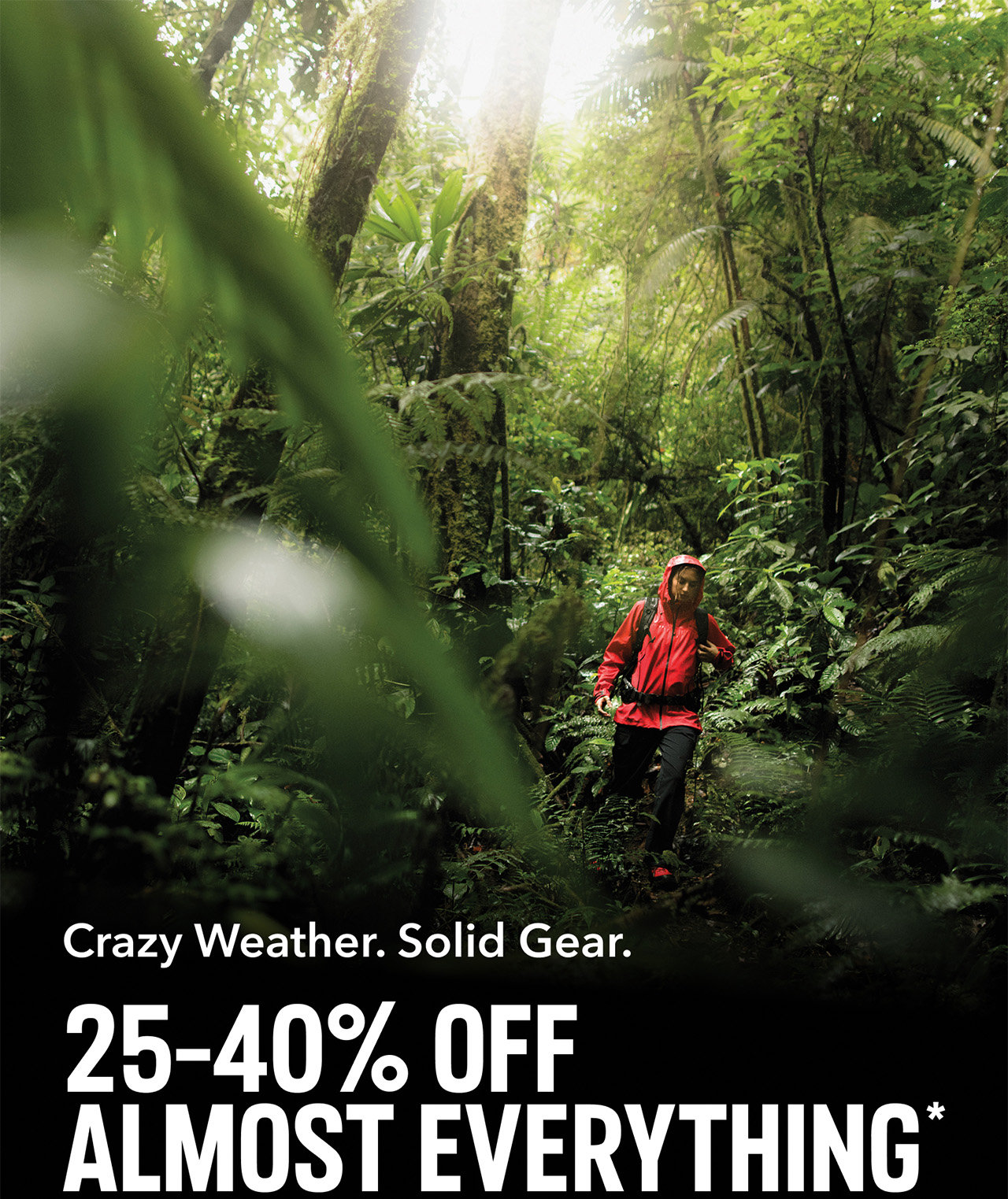 A hiker in a bright red rain jacket and black pants walking through a forest with text: 25-40% Off Almost Everything* CRAZY WEATHER. SOLID GEAR.