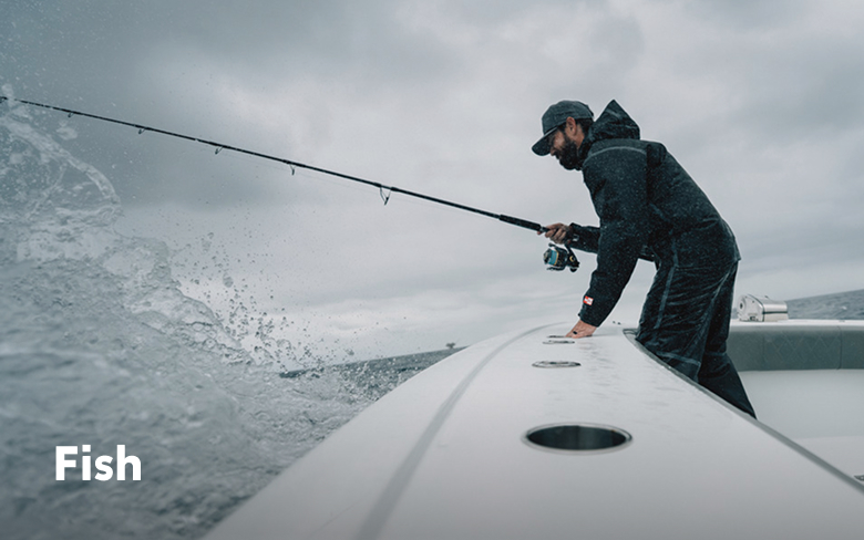 An angler on a boat, holding a rod, wearing a black jacket and pants with text: Fish