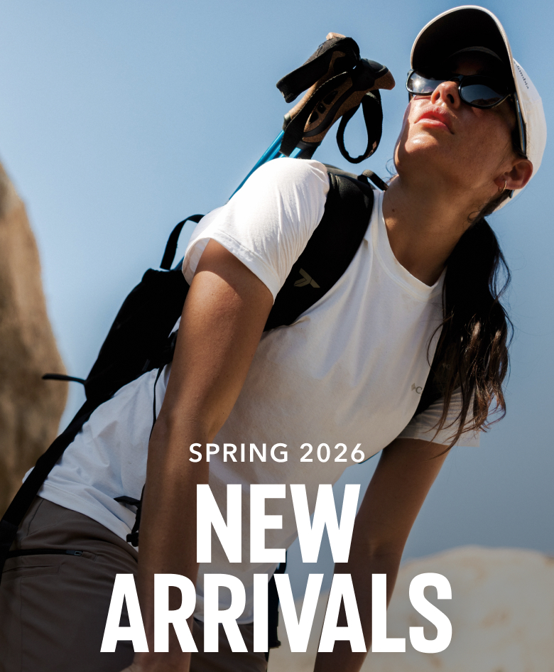 A woman in a white t-shirt, ball cap, and sunglasses, standing in a rocky outdoor setting wearing a black backpack with hiking poles strapped in with text: SPRING 2026 NEW ARRIVALS