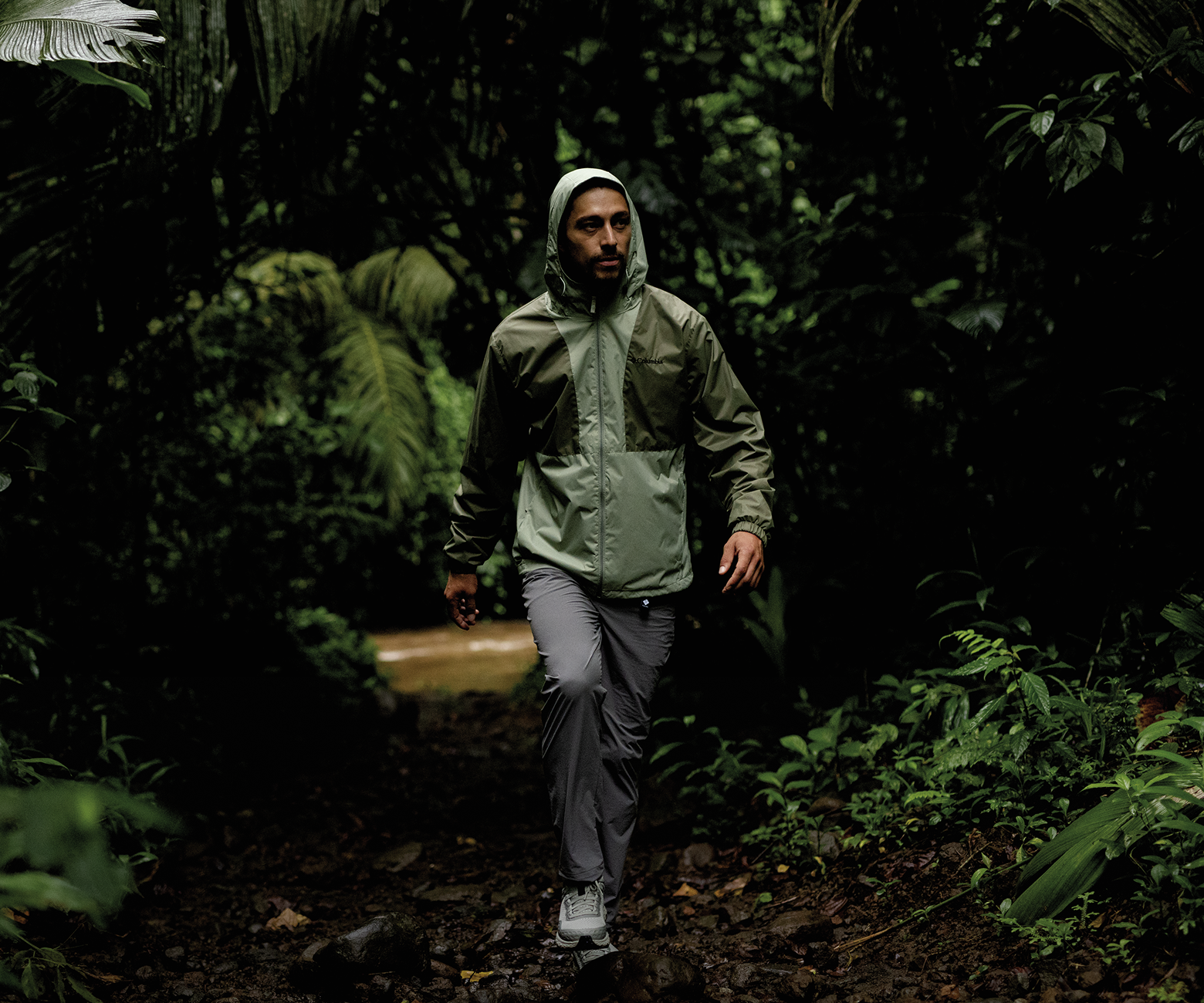 An image of a man walking on a trail through a forested area with his Columbia rain jacket on with his hood up.