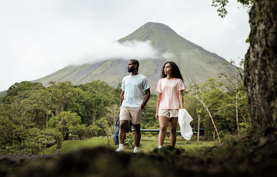 A man and woman walking together in a rainforest setting with a giant peak behind them.