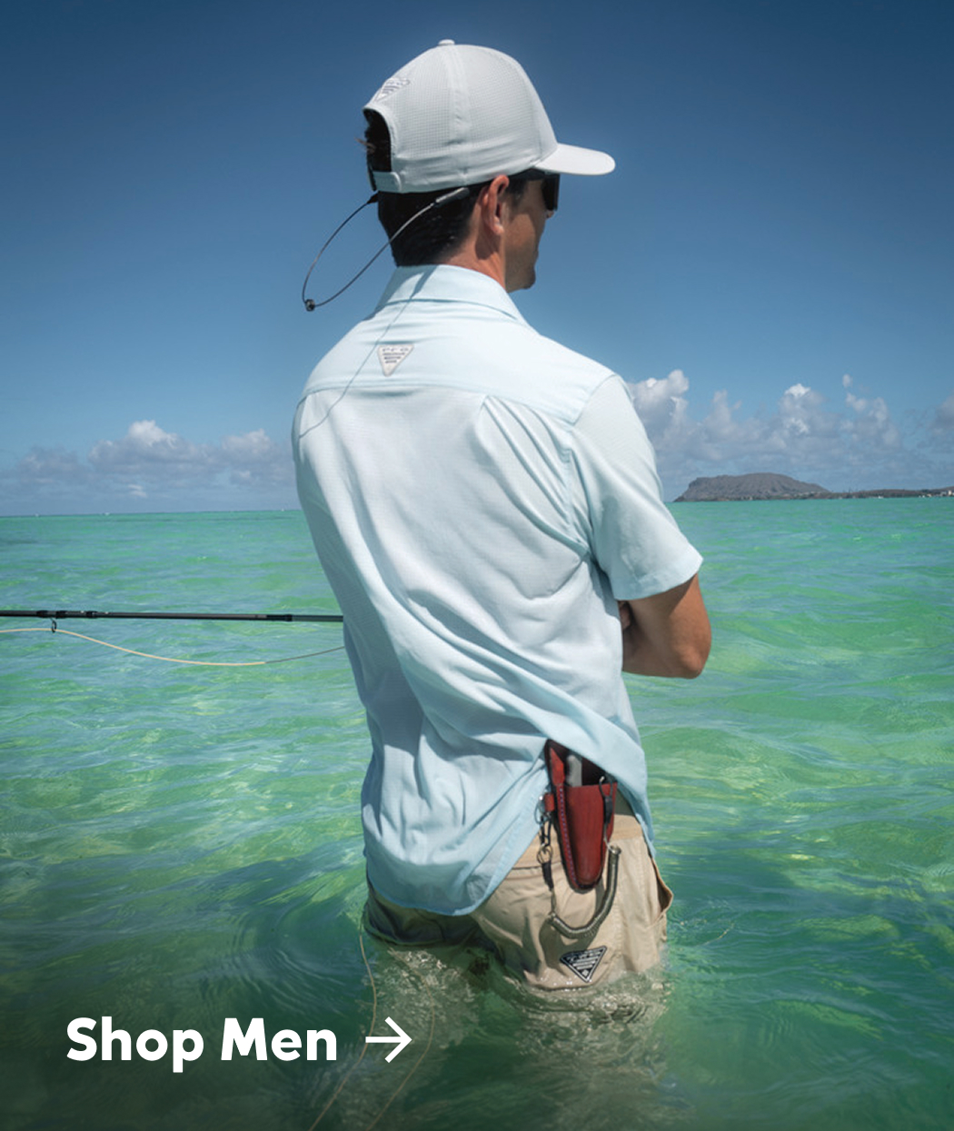 A man wearing a light blue short sleeve button-up, tan shorts, and a ball cap, facing away and holding a fishing rod as he stands in the water with text: Shop Men