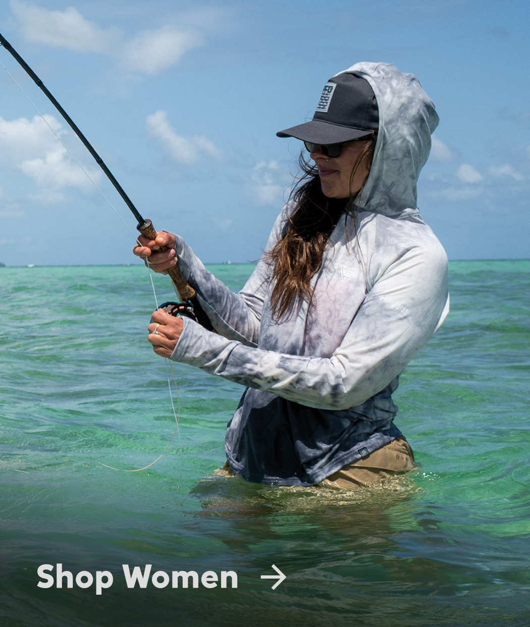 A woman wearing a blue sun hoodie with the hood up, and a blue ball cap and tan shorts, standing in the ocean with a fishing rod in hand with text: Shop Women