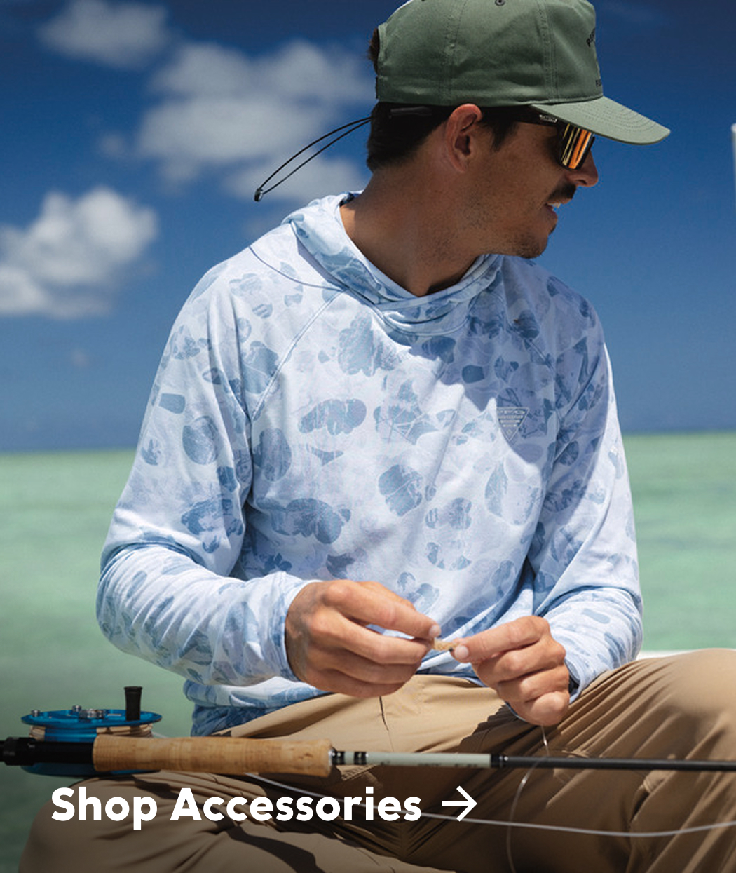 A man sitting on a boat, wearing a blue sun hoodie, tan pants, green ball cap, with a fishing rod balanced on his lap with text: Shop Accessories