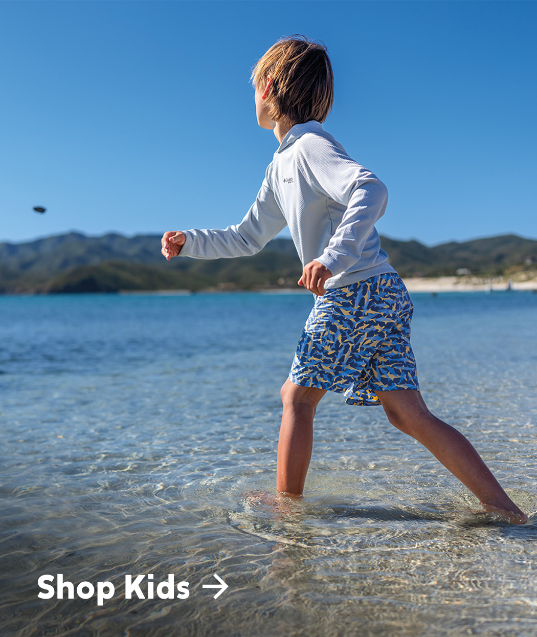 A little boy running along the shore, wearing a blue sun hoodie, blue swim trunks with text: Shop Kids