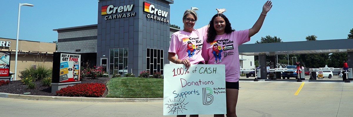 Two volunteers holding a BBBS sign in front of Crew Carwash