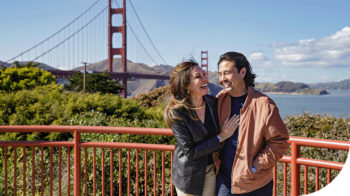 A man and woman are standing together next to a bridge outdoors. The sky is partly cloudy, and a body of water is visible nearby. They are dressed casually, and there is a fence in the foreground with buildings in the background.
