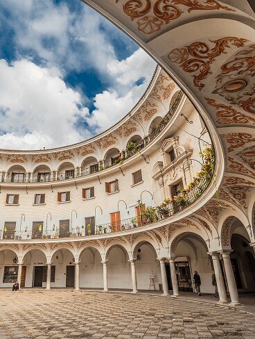 Oval-shaped courtyard with arched walkways, ornate painted ceiling, balconies, and blue sky with clouds above. A few people are visible in the scene.