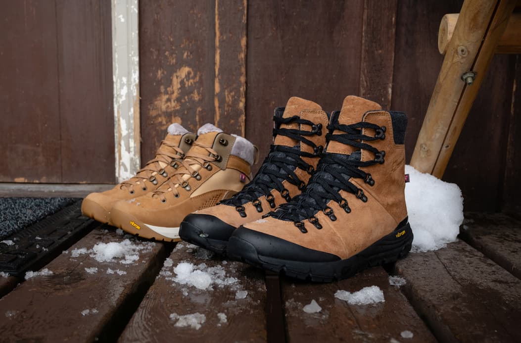 Women’s Inquire Winter and men’s Arctic 600 Side-Zip boots sitting side by side on a snowy wooden porch, showcasing their weather-ready uppers and Vibram outsoles.