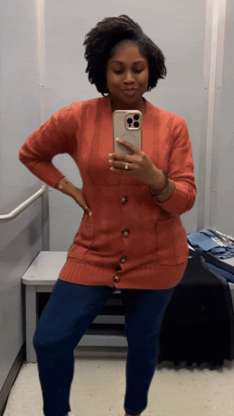 GIF of a person in a fitting room wearing a black top with green floral pants and an orange button up cardigan with blue jeans.. 