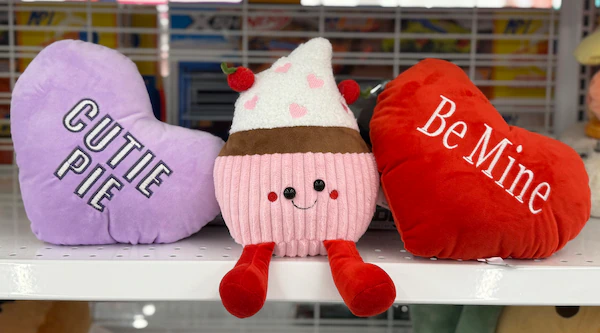 A display of three plush toys including a purple heart with “Cutie Pie,” a pink cupcake plush with dangling red legs and strawberry decorations, and a red heart with “Be Mine.”