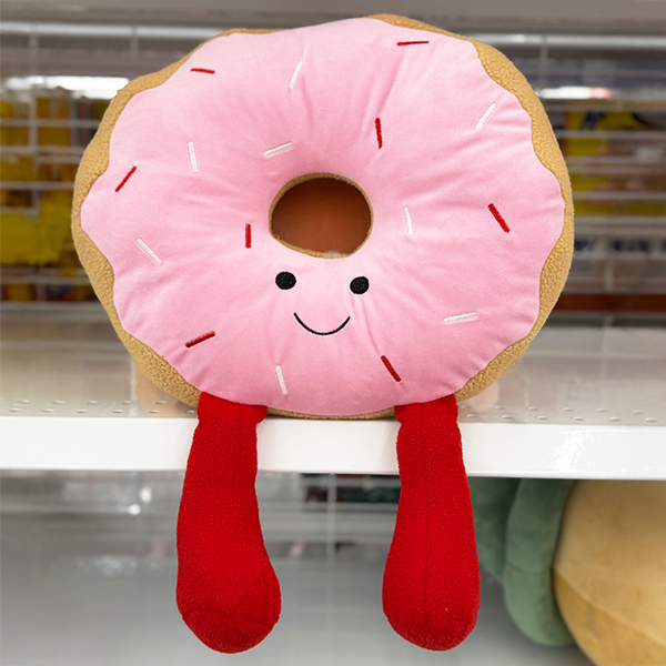 A round donut plush toy with light pink frosting, stitched sprinkles, a smiling face, and two long red plush legs hanging down.