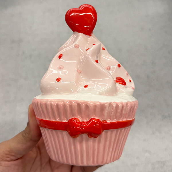A ceramic cupcake figurine with ribbed pink sides, white and pale pink swirled frosting, red accents, and a shiny red heart on top.