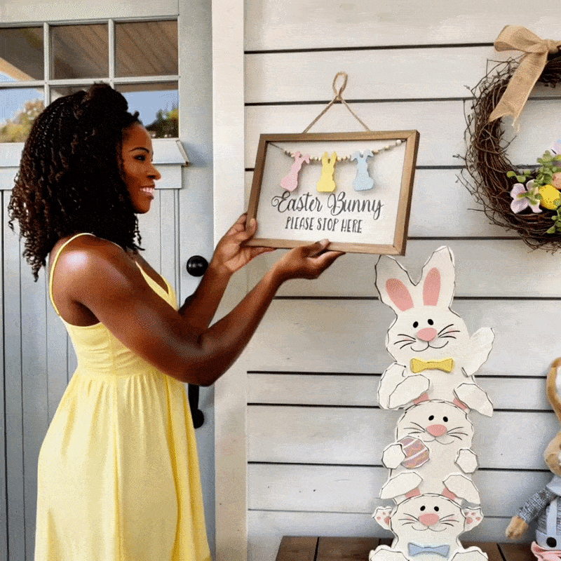 A person wearing a yellow dress holds a framed Easter sign on a white porch wall decorated with a bunny stack decoration and a floral wreath.