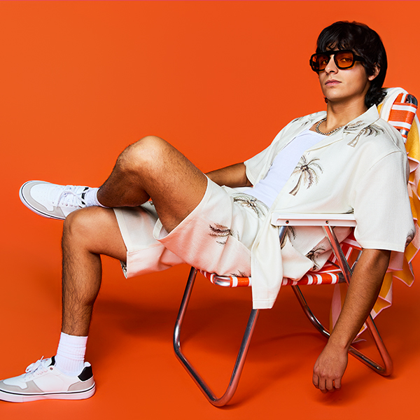 A person wearing a light short-sleeve shirt and matching shorts sits in a striped lounge chair, styled with sunglasses and white sneakers against a bold orange backdrop.
