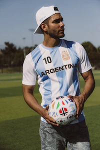 A person in a light blue and white striped Argentina jersey holds a soccer ball while standing on a grassy field.